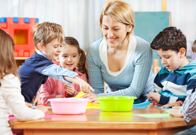 Afbeelding van juf die met 4 kinderen aan een tafel zit met 2 bakjes en gekleurd papier